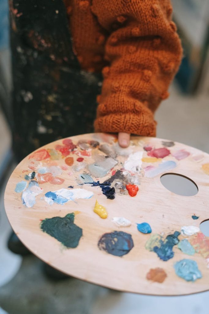 blue-and-white-round-ornament-6816527 A child in a cozy sweater holding a colorful paint palette during art class.